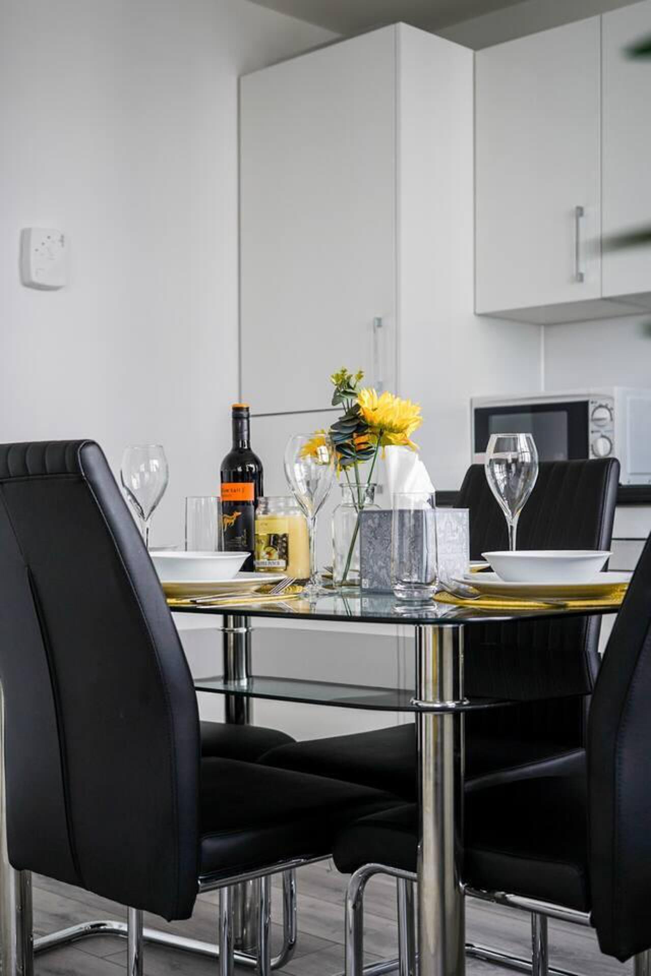 Een moderne keuken met een eettafel gedekt voor twee personen, met zwarte stoelen, een glazen tafel en een vaas met gele bloemen.