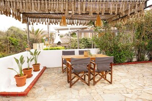 Eine überdachte Terrasse mit Steinboden, einem Holztisch mit Stühlen und Topfpflanzen.