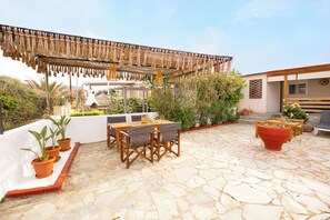 Eine Terrasse im Freien mit Steinboden, Holztisch und -stühlen, Kübelpflanzen und einer Pergola mit hängenden Lichtern.