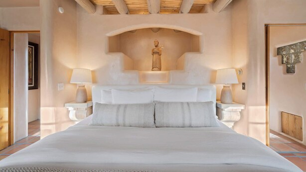 A bedroom with a large bed, two bedside tables with lamps, a wooden ceiling, and a statue on the wall. - Luxurious Renovated Pueblo Style Home (Santa Fe)