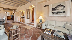 A spacious living area with a stone floor, a wooden dining table, a sofa, and a coffee table. There is a kitchen in the background with wooden cabinets and a hanging light fixture. - Luxurious Renovated Pueblo Style Home (Santa Fe)