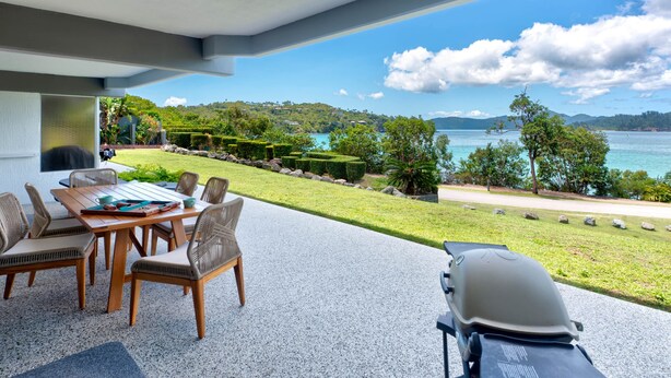 A patio with a table and chairs, a barbecue grill, and a view of a lake and hills. - Frangipani Lodge F7, Frangipani Lodge F7 (Hamilton Island)