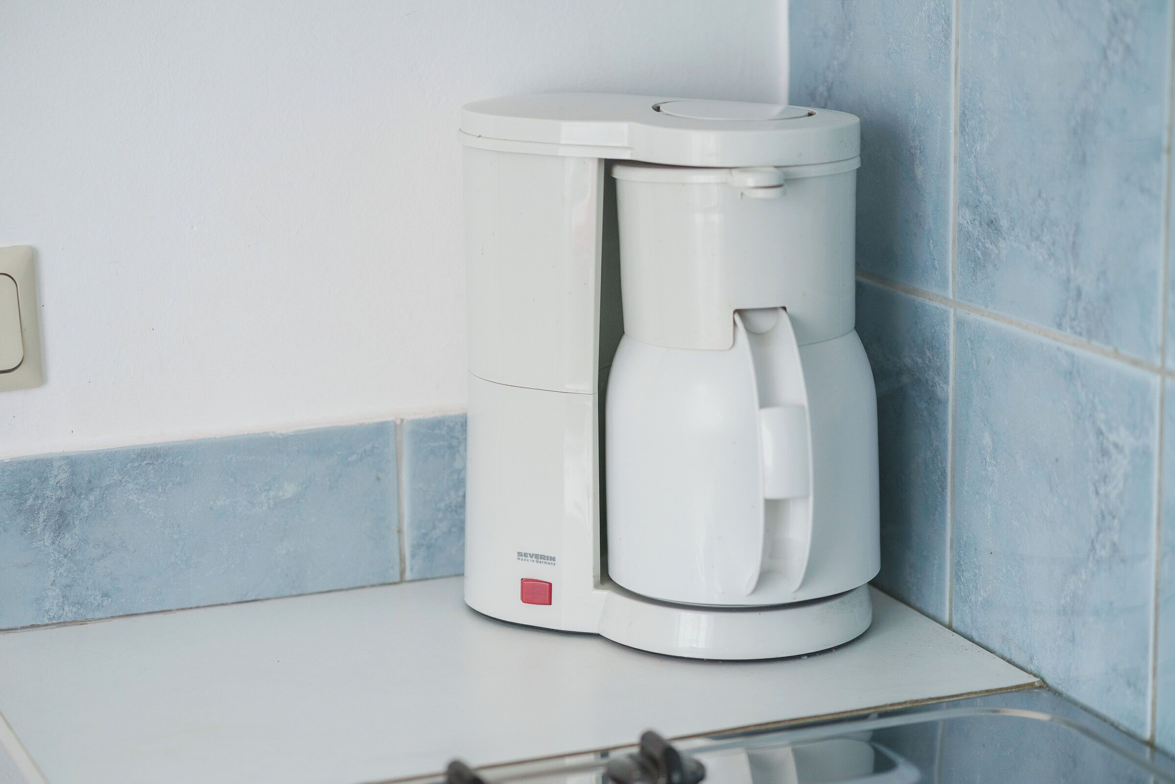 A white coffee maker on a white countertop with blue tiles in the background.