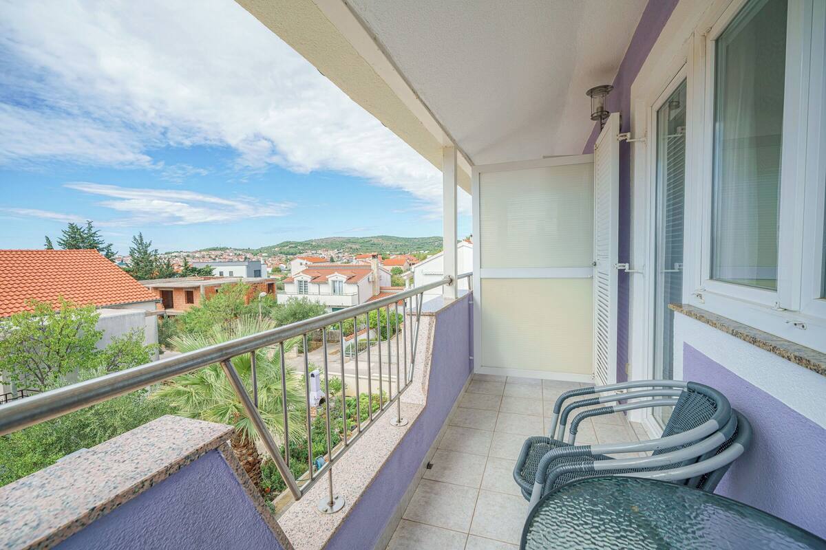 Un balcon doté d’une balustrade en métal, de deux fauteuils en osier et offrant une vue sur une ville aux bâtiments aux toits rouges.