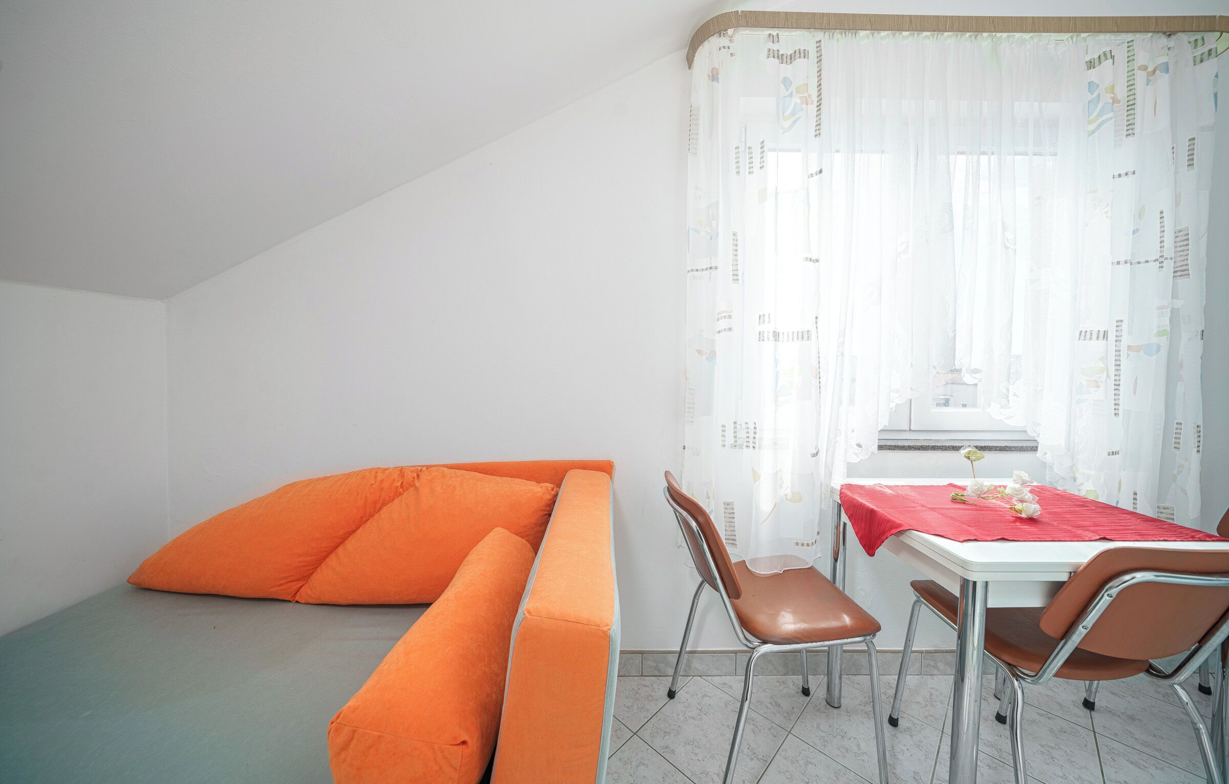 A room with a white wall, a white ceiling, a white window with a white curtain, a white table with a red tablecloth, and a brown sofa with orange cushions.