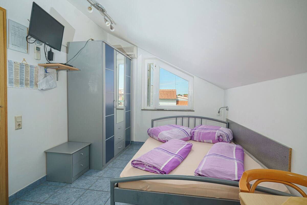 Une chambre à coucher de taille modeste, comprenant un lit, un bureau, une chaise, une armoire, un téléviseur et une fenêtre donnant sur des immeubles.