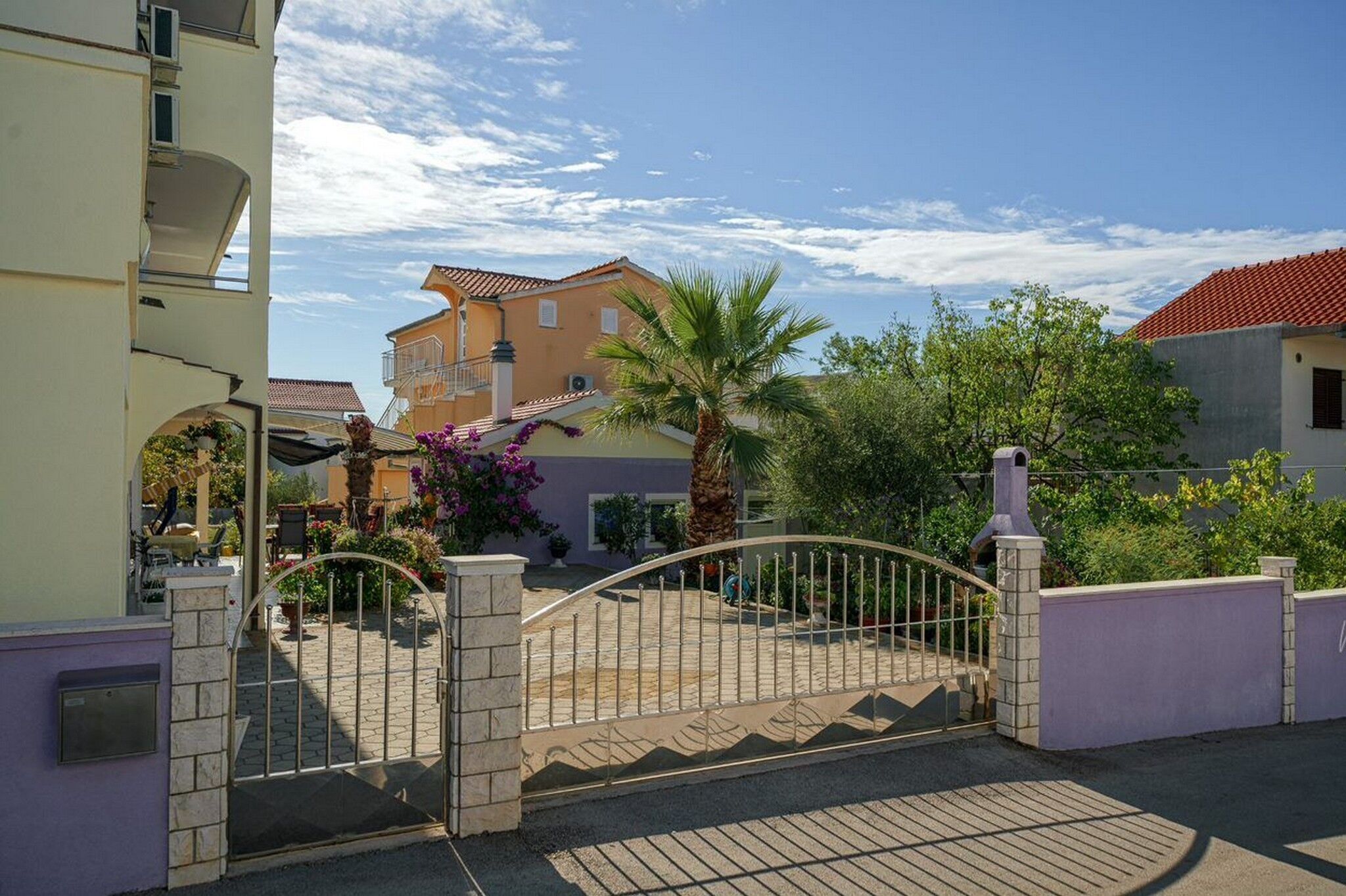 A gated entrance with a palm tree and a building with a red-tiled roof in the background.