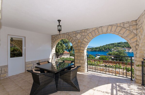 Une terrasse couverte avec une arche en pierre, une table et des chaises, et une vue sur une étendue d’eau bleue et une végétation luxuriante.