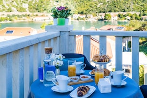 Mesa de pequeno-almoço com croissants, sumo de laranja e chávenas de café numa varanda com vista para um lago.
