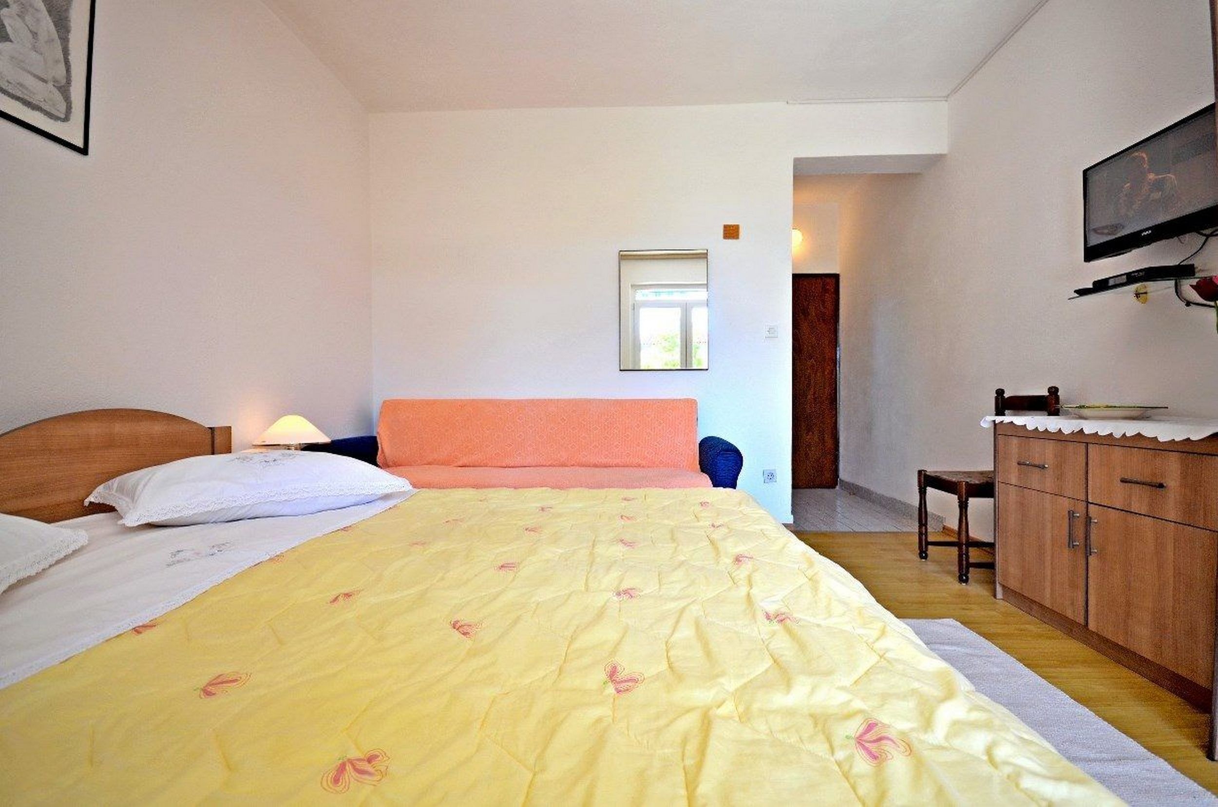 Une chambre à coucher comprenant un lit, une tête de lit en bois, une table de chevet, un téléviseur fixé au mur, une petite table et un meuble de rangement.