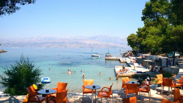 Vista sulla costa con acque limpide, imbarcazioni e persone che nuotano. Sulla spiaggia ci sono sedie e tavoli arancioni.