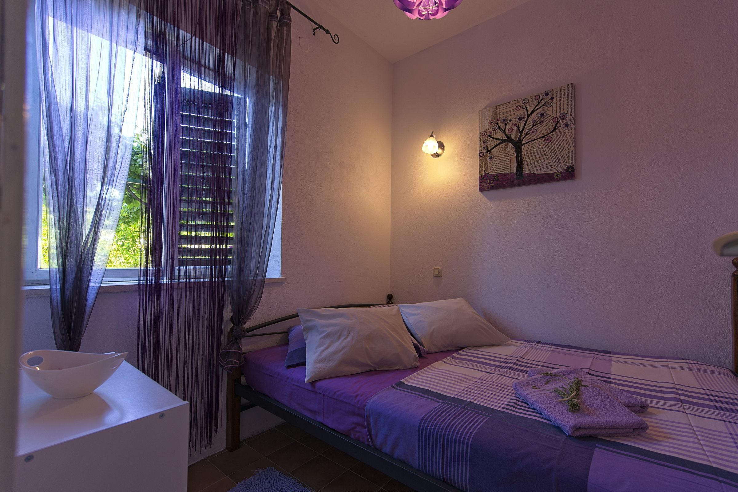 A bedroom with a bed, a bedside table, a window with curtains, a wall-mounted light, and a painting.