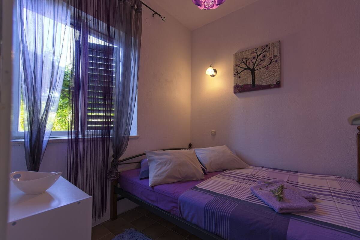 Ein Schlafzimmer mit einem Bett, einem Nachttisch, einem Fenster mit Vorhängen, einer Wandleuchte und einem Gemälde.