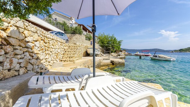 Eine Steinsicht, weiße Lounge-Stühle und ein klares, blaues Meer mit Booten.
