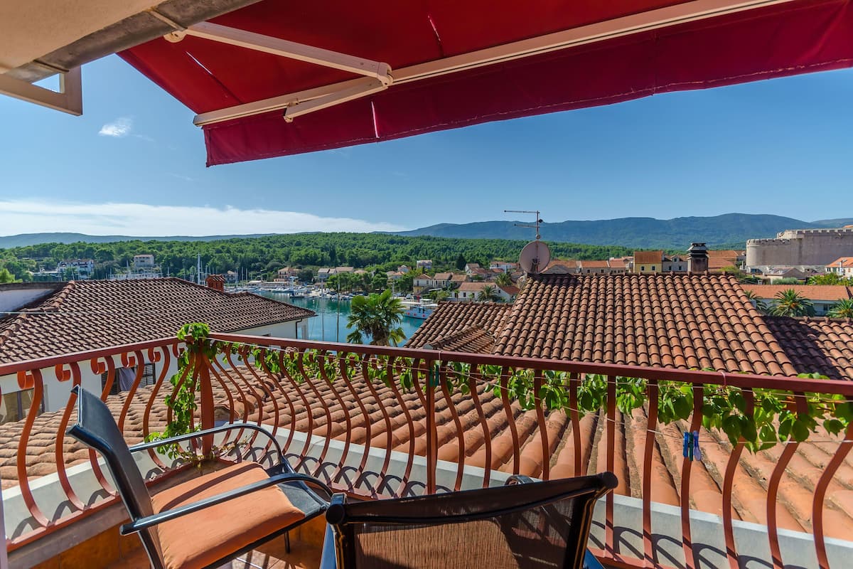 Un balcon avec un parasol rouge, une chaise et une vue sur une ville, avec des bâtiments et des montagnes en arrière-plan.