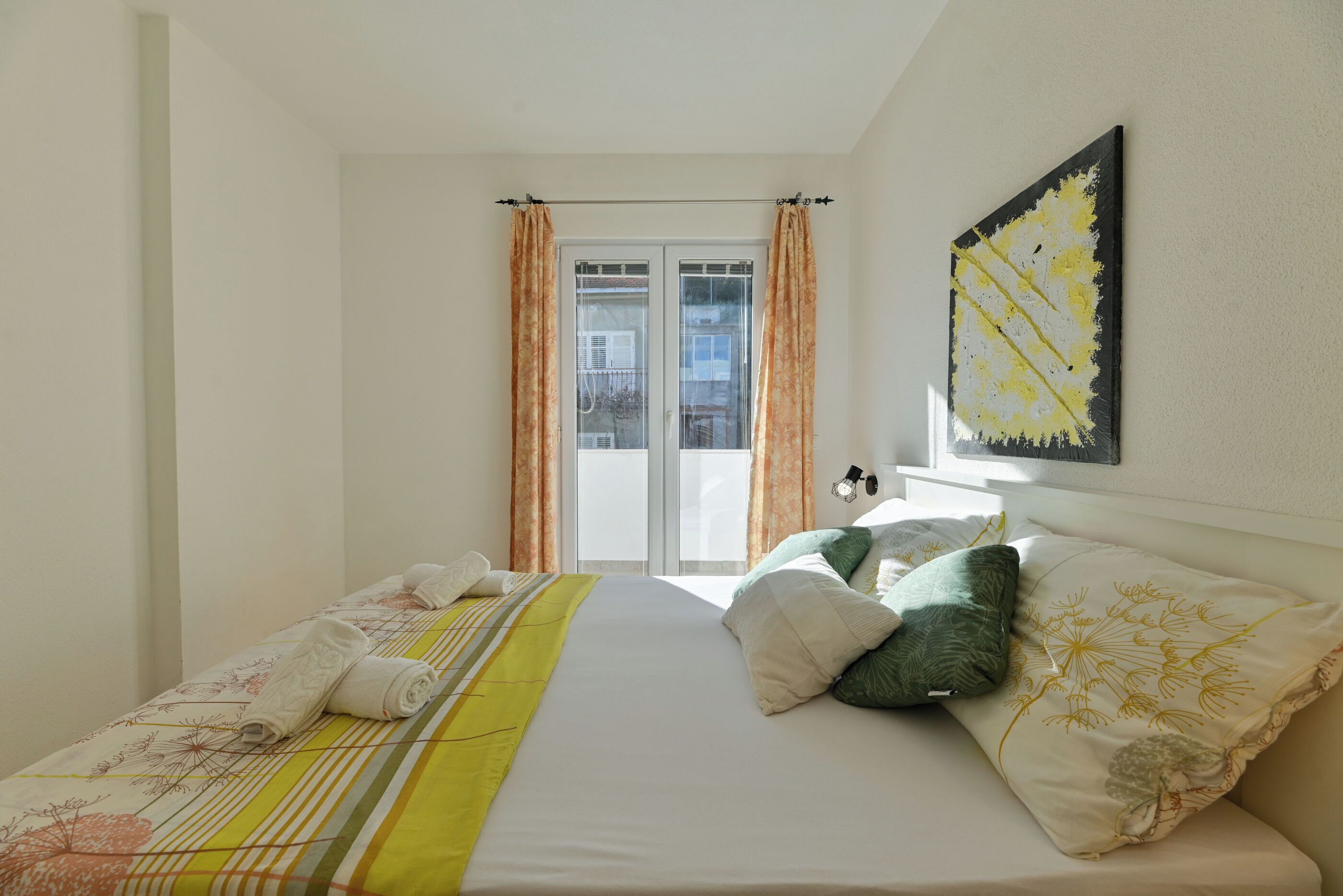 Een slaapkamer met een bed, kussens, een raam met gordijnen en een kunstwerk aan de muur.