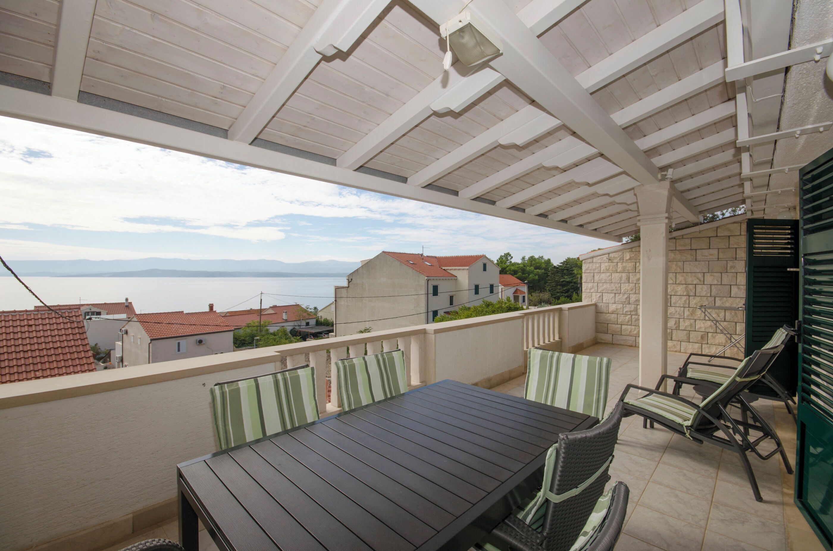 A covered outdoor patio with a table and chairs, overlooking a residential area with houses and a view of the sea.