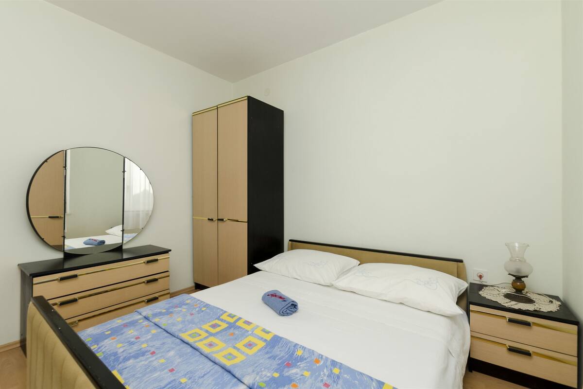 A bedroom with a bed, a bedside table with a lamp, a dresser, a wardrobe, and a round mirror.