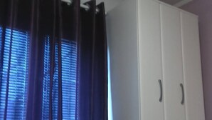 Una habitación con un armario blanco, un estante con libros y una planta pequeña, y una ventana con cortinas azules.