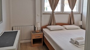 A bedroom with a bed, two bedside lamps, a nightstand, a radiator, and beige curtains. - Apartments Natasa (Supetar)