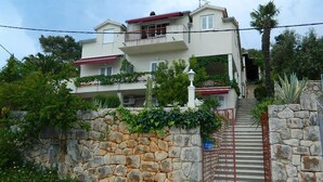 A two-story house with a stone wall, a red gate, and a staircase leading to the entrance. - Apartment and Room Darinka (Vrboska)