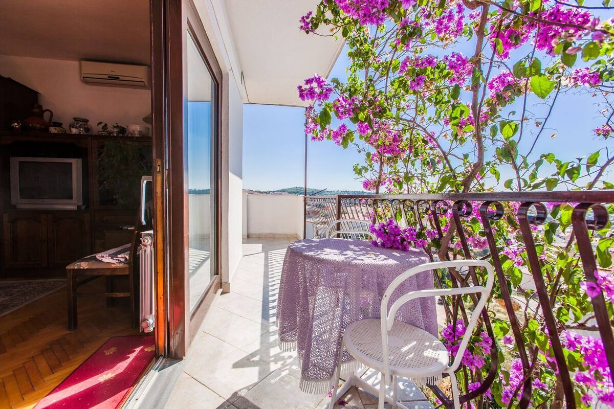 Un balcón con una mesa, sillas, un televisor y una planta con flores púrpuras.