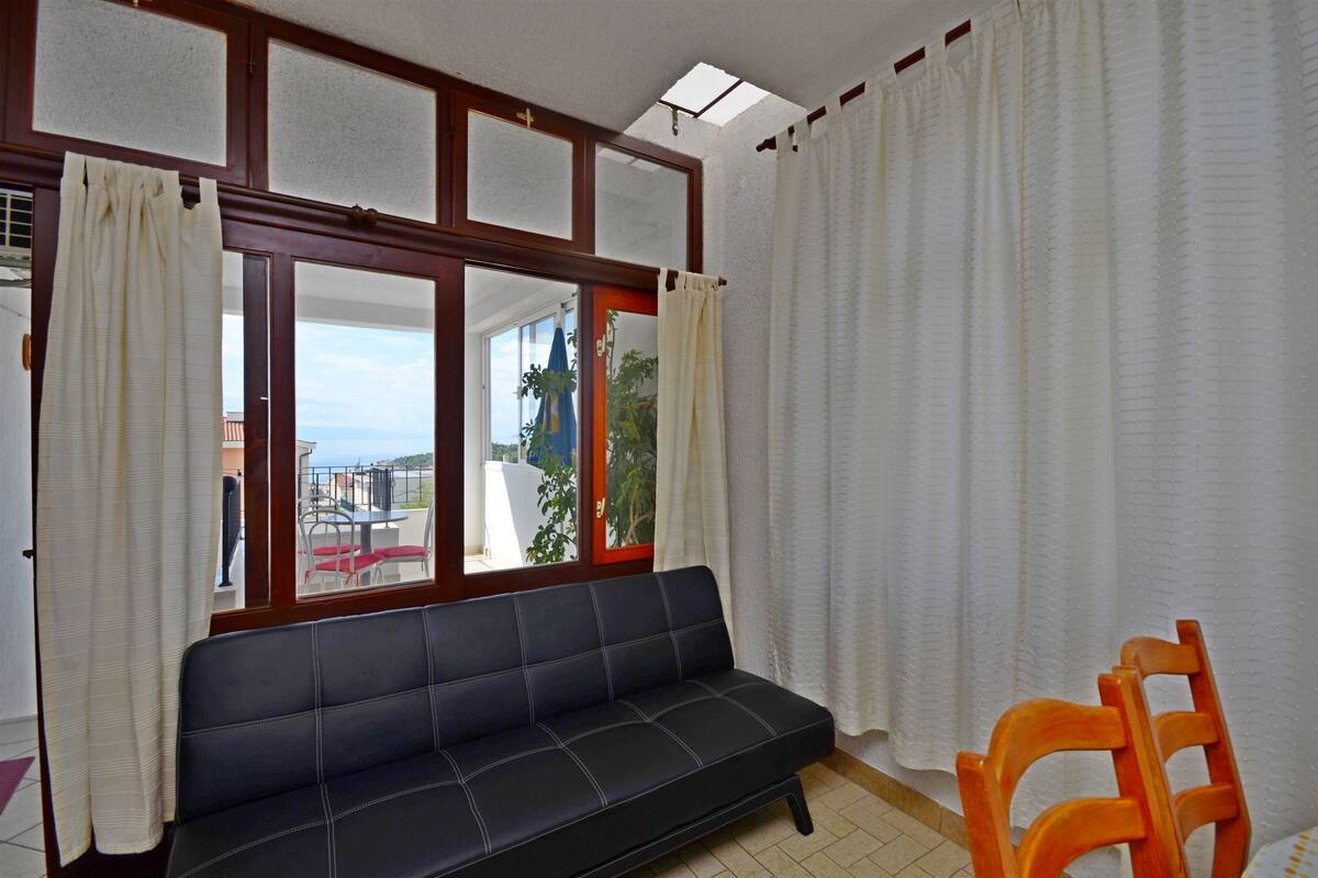 Una habitación con un sofá oscuro, una silla de madera, una ventana con cortinas y vistas al exterior.