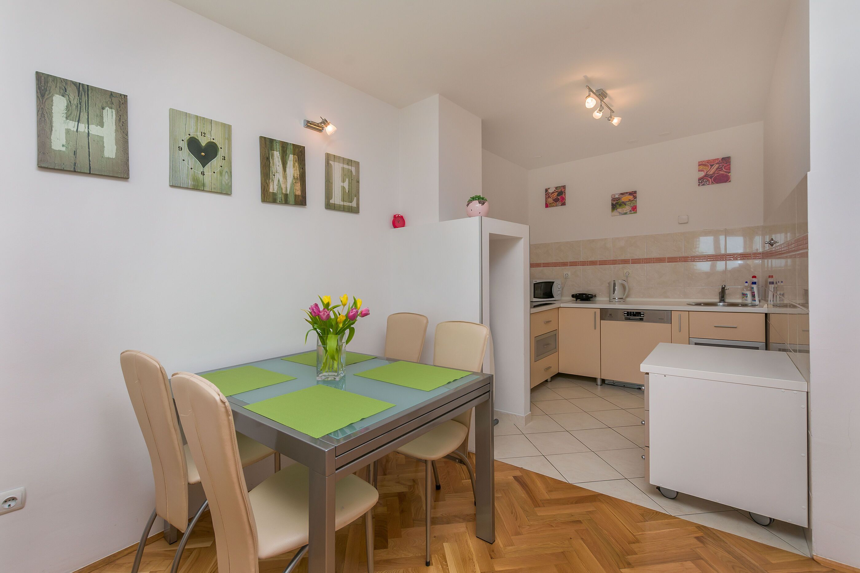 A dining area with a table, chairs, and a vase of flowers. There is a kitchen in the background with cabinets and appliances.