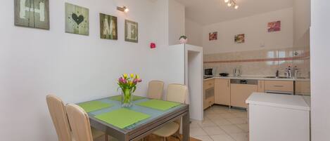 A dining area with a table, chairs, and a vase of flowers. There is a kitchen in the background with cabinets and appliances.