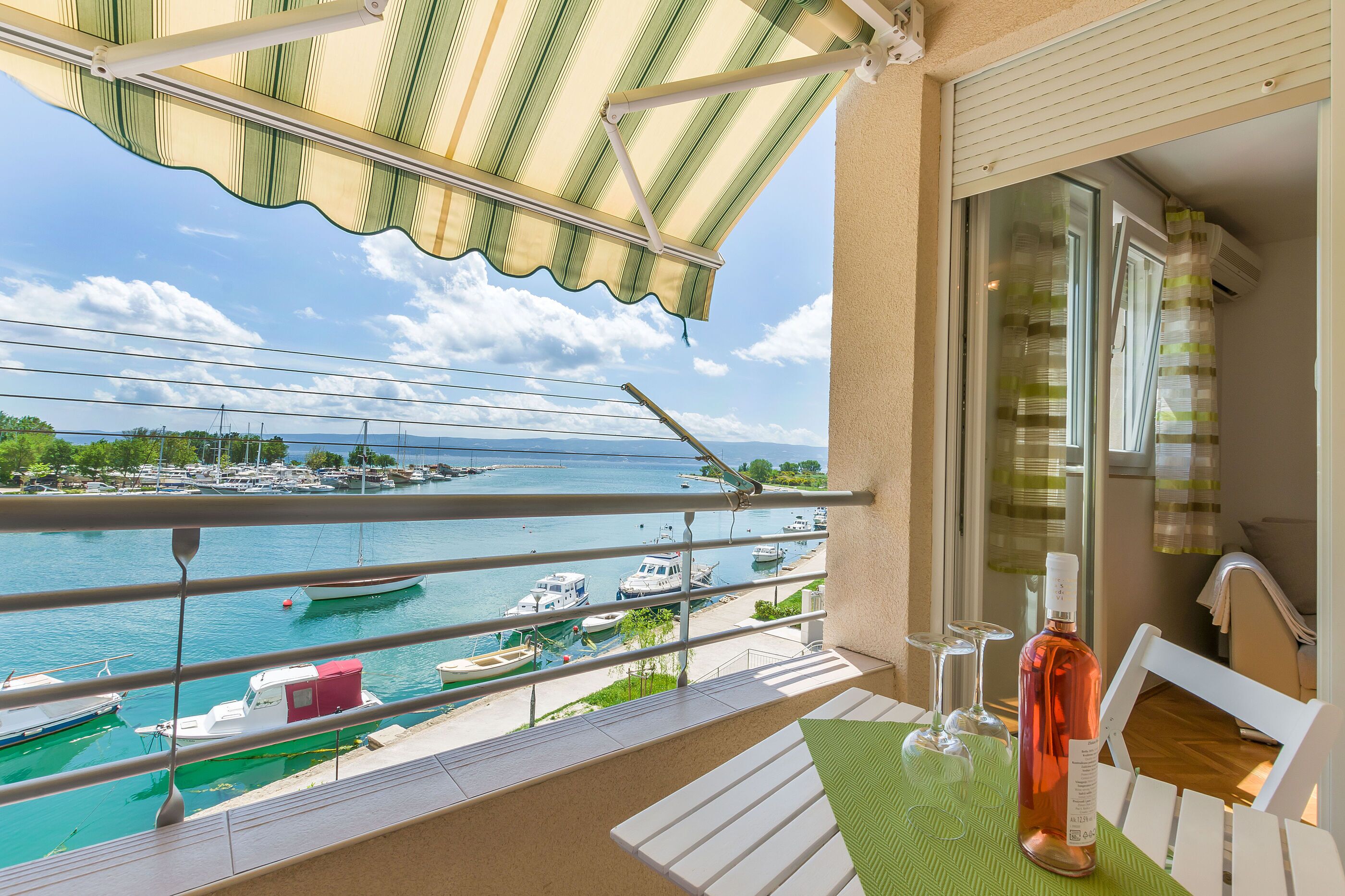 A balcony with a view of a marina, a bottle of wine, and two glasses on a table.