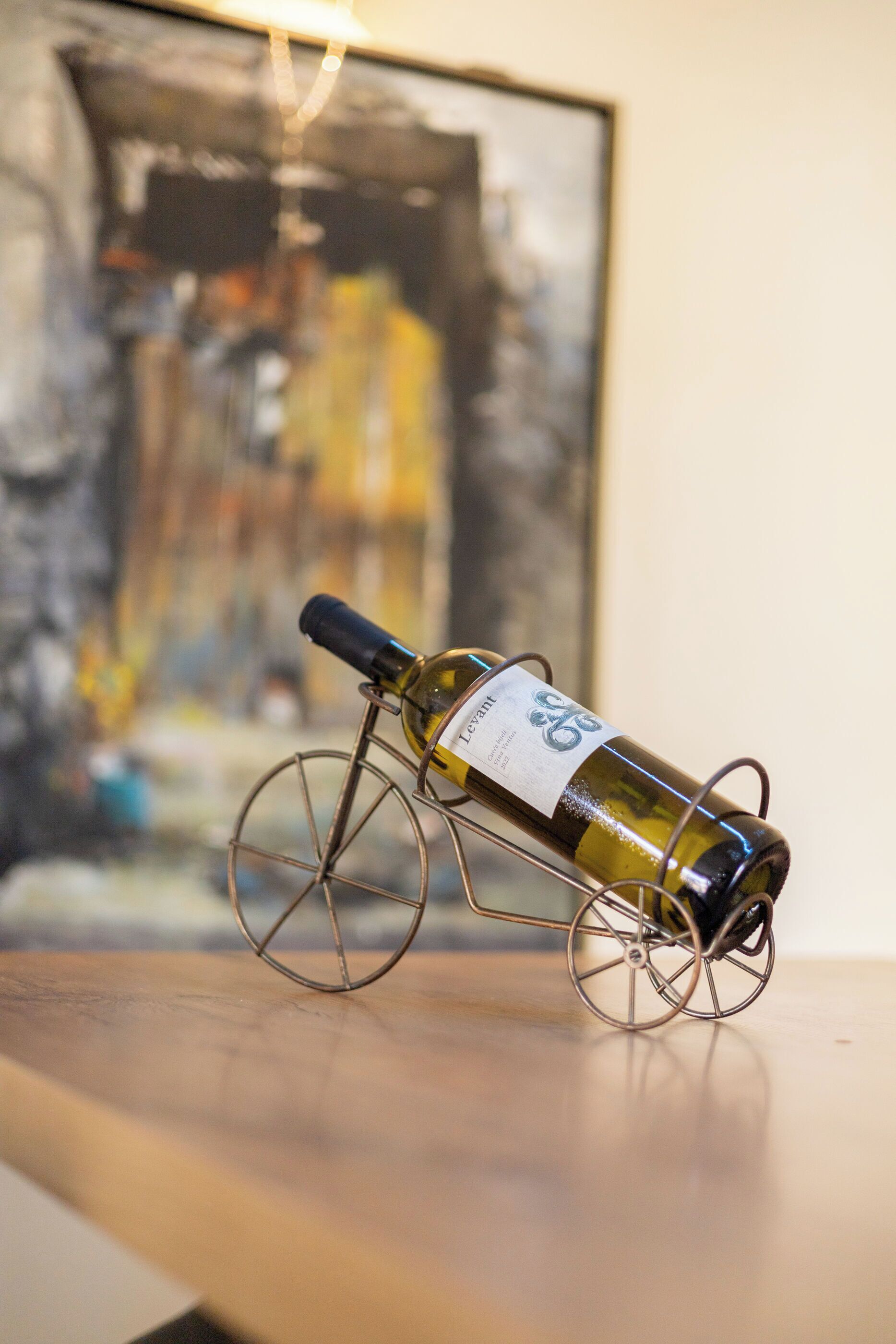 A wine bottle on a metal stand with a label, placed on a wooden surface with a blurred painting in the background.