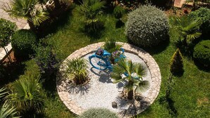 Uma área de jardim circular com uma borda de pedra, rodeada por diversos tipos de plantas e uma bicicleta azul.