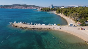 Vista aérea de una zona costera con playa de arena, barcos y un paisaje urbano al fondo.