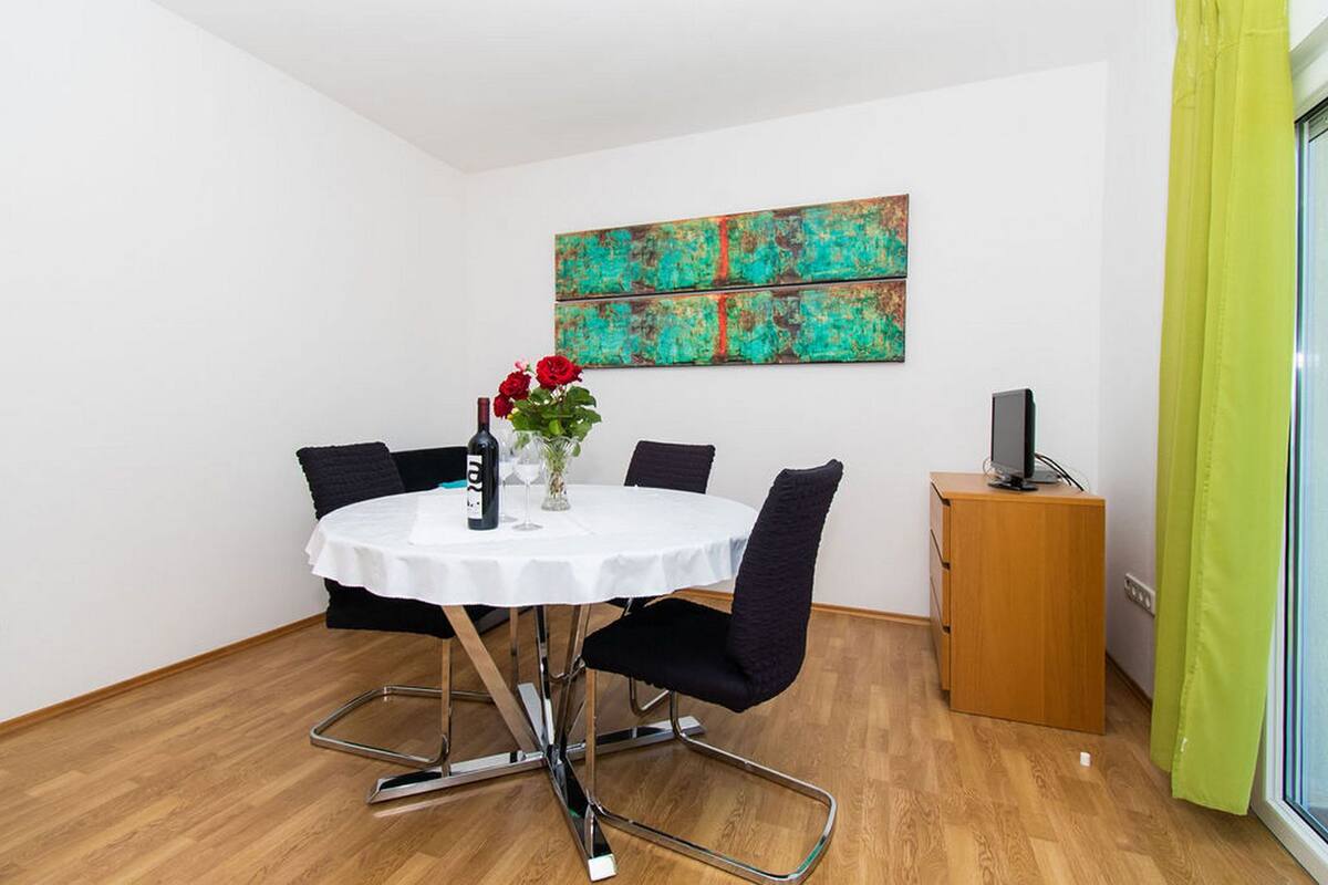 A round table with a white tablecloth, six black chairs, a bottle of wine, and a vase with red flowers. A colorful abstract painting is on the wall above the table.