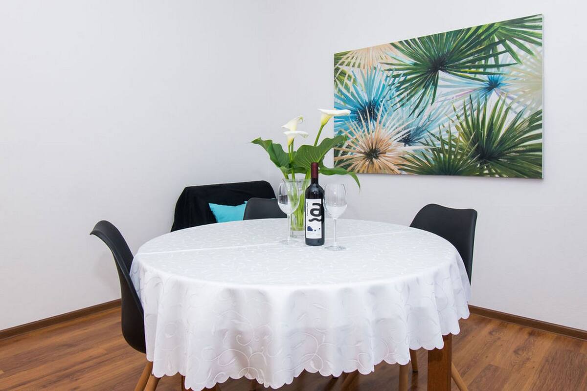 A round dining table with a white tablecloth, a vase with flowers, a wine bottle, and a glass. There is a painting on the wall behind the table.