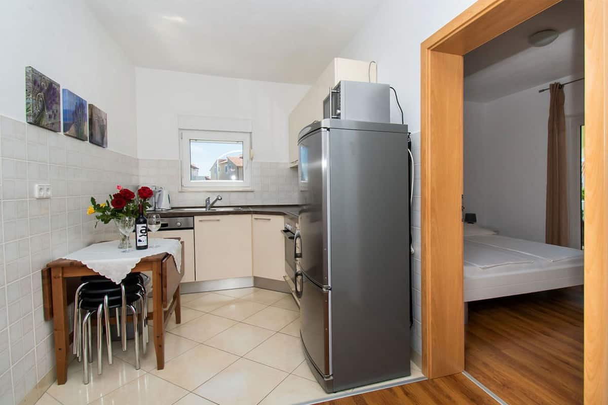A compact kitchen with a refrigerator, a dining table with chairs, a small window, and a wooden door leading to a bedroom.