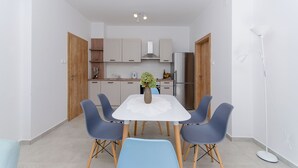 A modern kitchen with white cabinets, a central island, and a dining area with a white table and blue chairs. - Apartments Koras (Kneza)