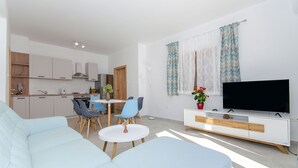 A modern living room with a sofa, a dining table with chairs, a TV on a white cabinet, and a kitchen in the background. - Apartments Koras (Kneza)