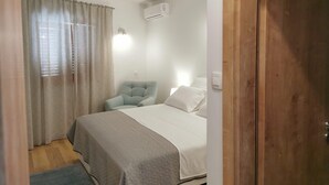 A bedroom with a bed, a chair, a window with curtains, an air conditioning unit, and a wooden door. - Apartments Koras (Kneza)