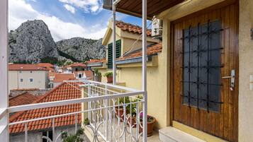 A balcony with a white railing, a wooden door with a metal grid, and terracotta-tiled roofs in the background.