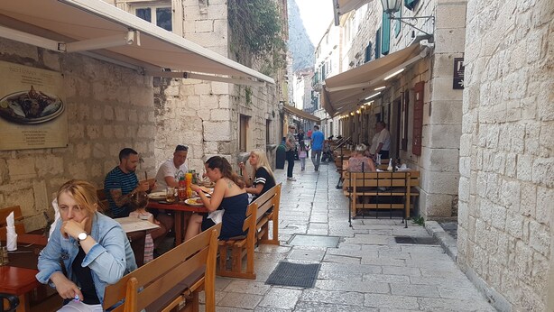 Una calle estrecha con mesas en terrazas, edificios de piedra y un letrero de restaurante.