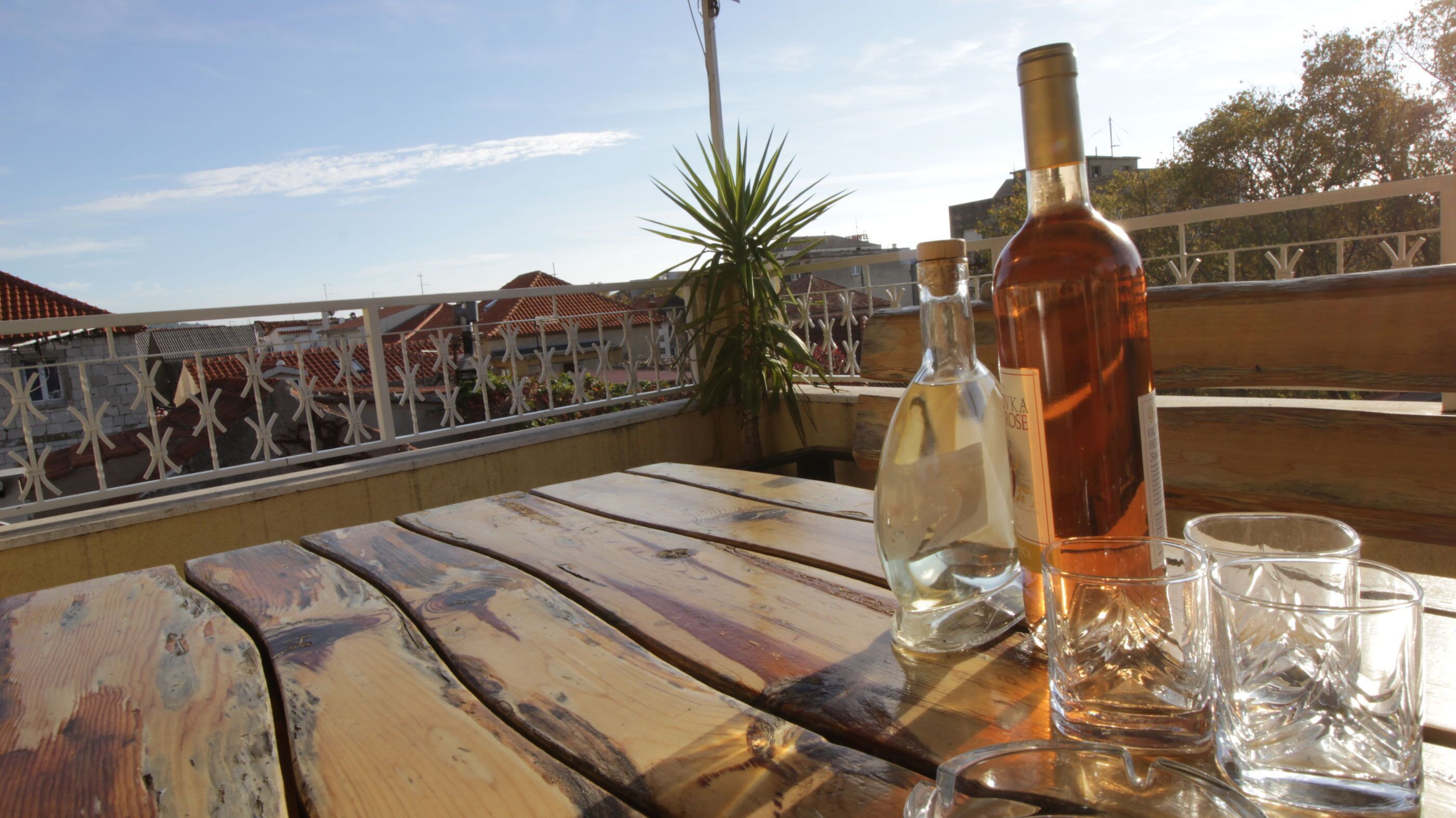 A wooden table with bottles and glasses on a balcony with a railing and a view of buildings.
