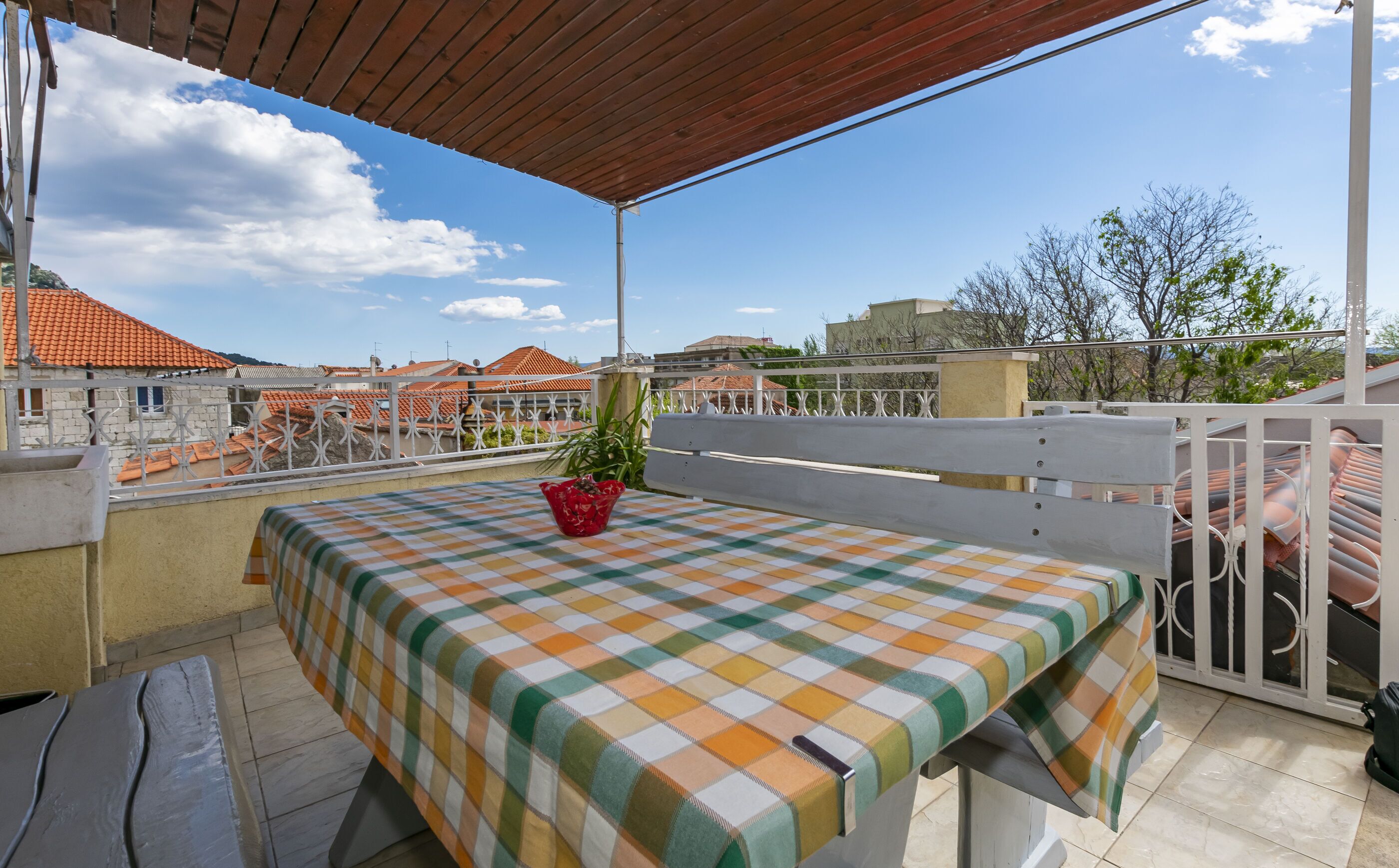 A balcony with a checkered tablecloth, a potted plant, and a view of buildings with orange roofs.