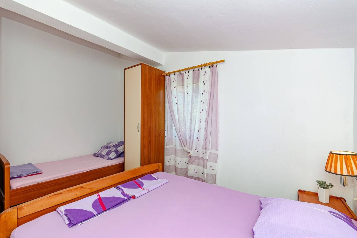A bedroom with two beds, a wooden wardrobe, a window with sheer curtains, and a bedside lamp.