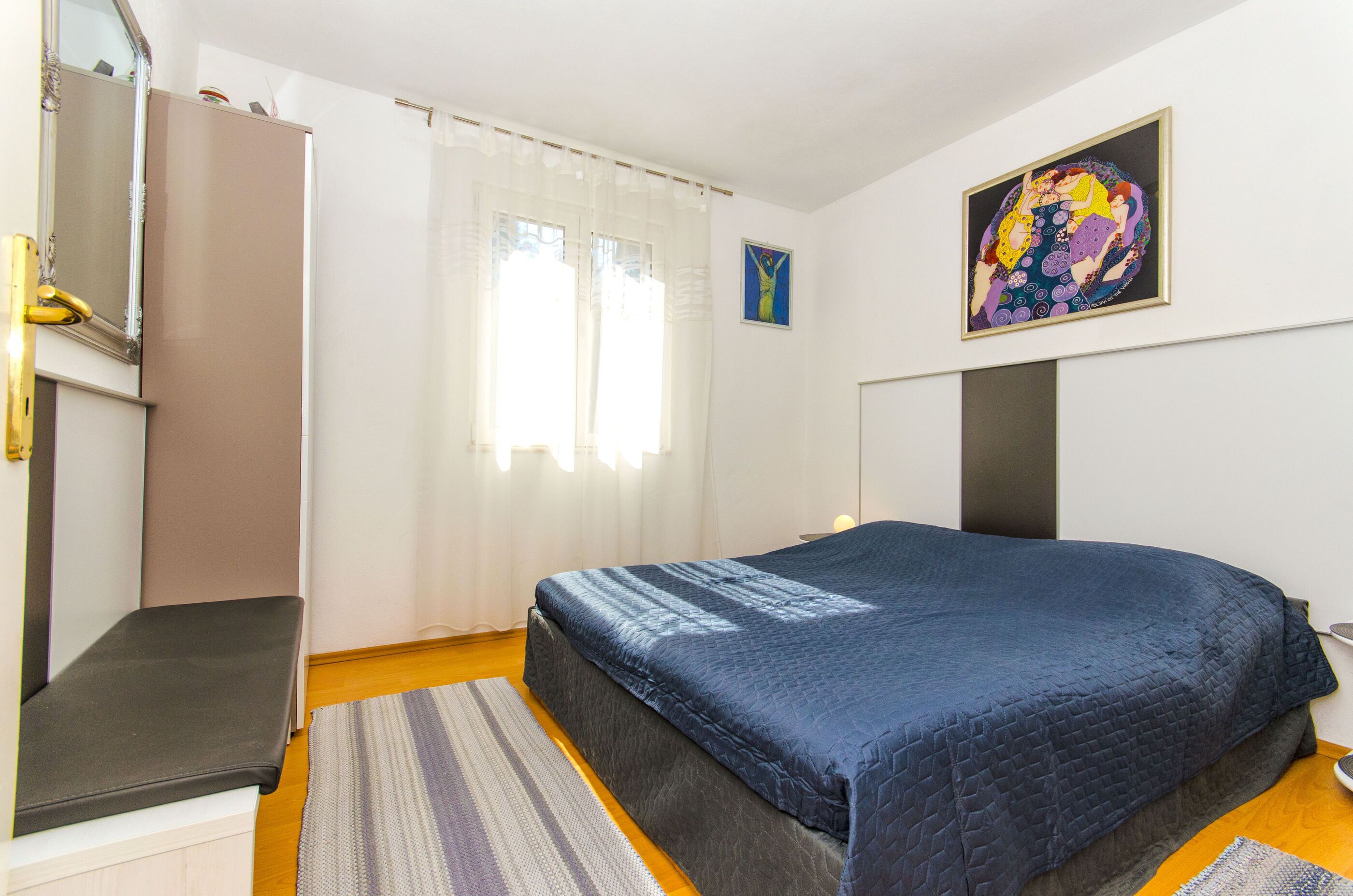 A bedroom with a bed, a bench, a wardrobe, a painting, and a window with curtains.