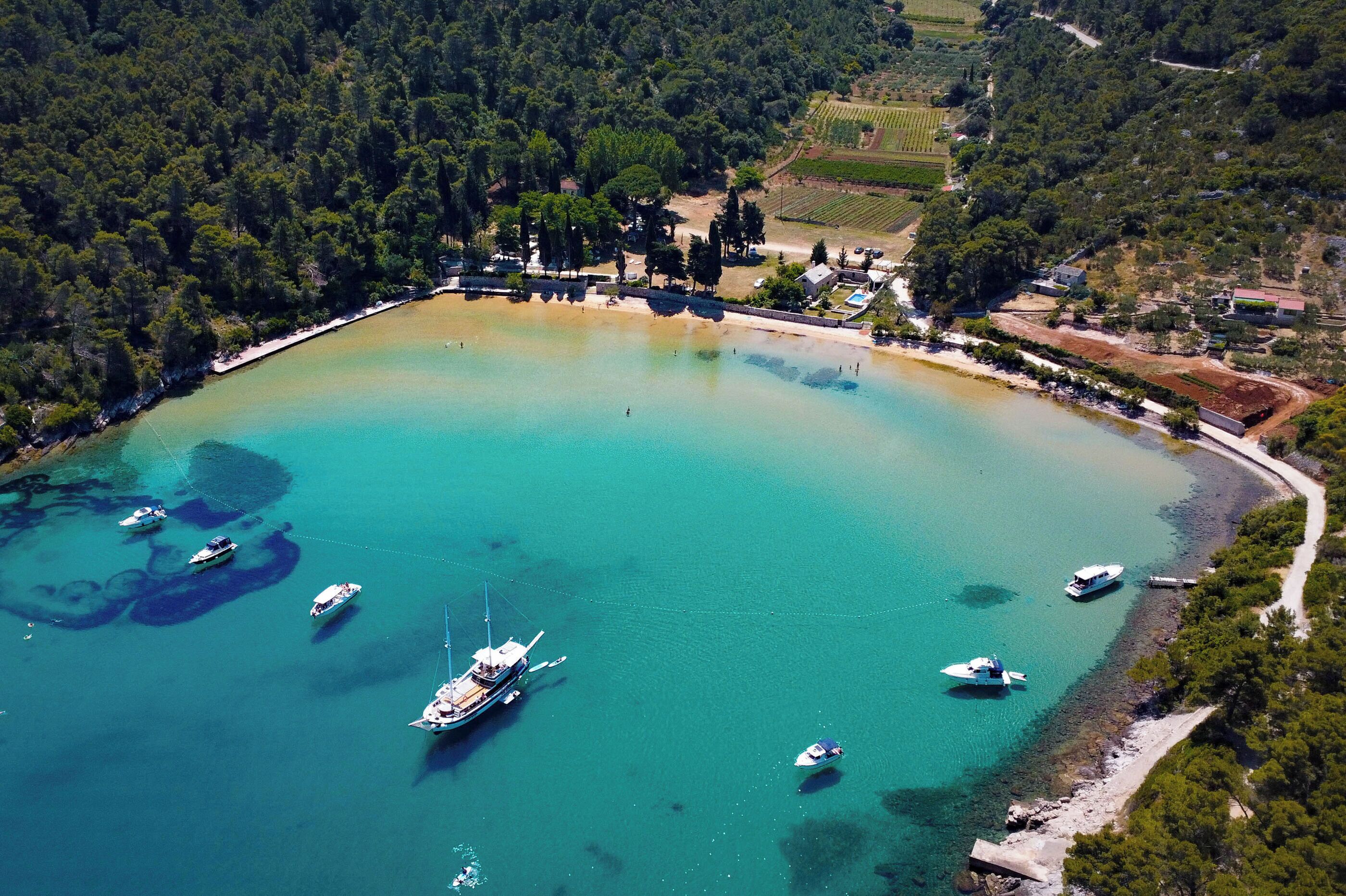 An aerial view of a coastal area with boats docked near a beach, surrounded by dense forest and agricultural fields.