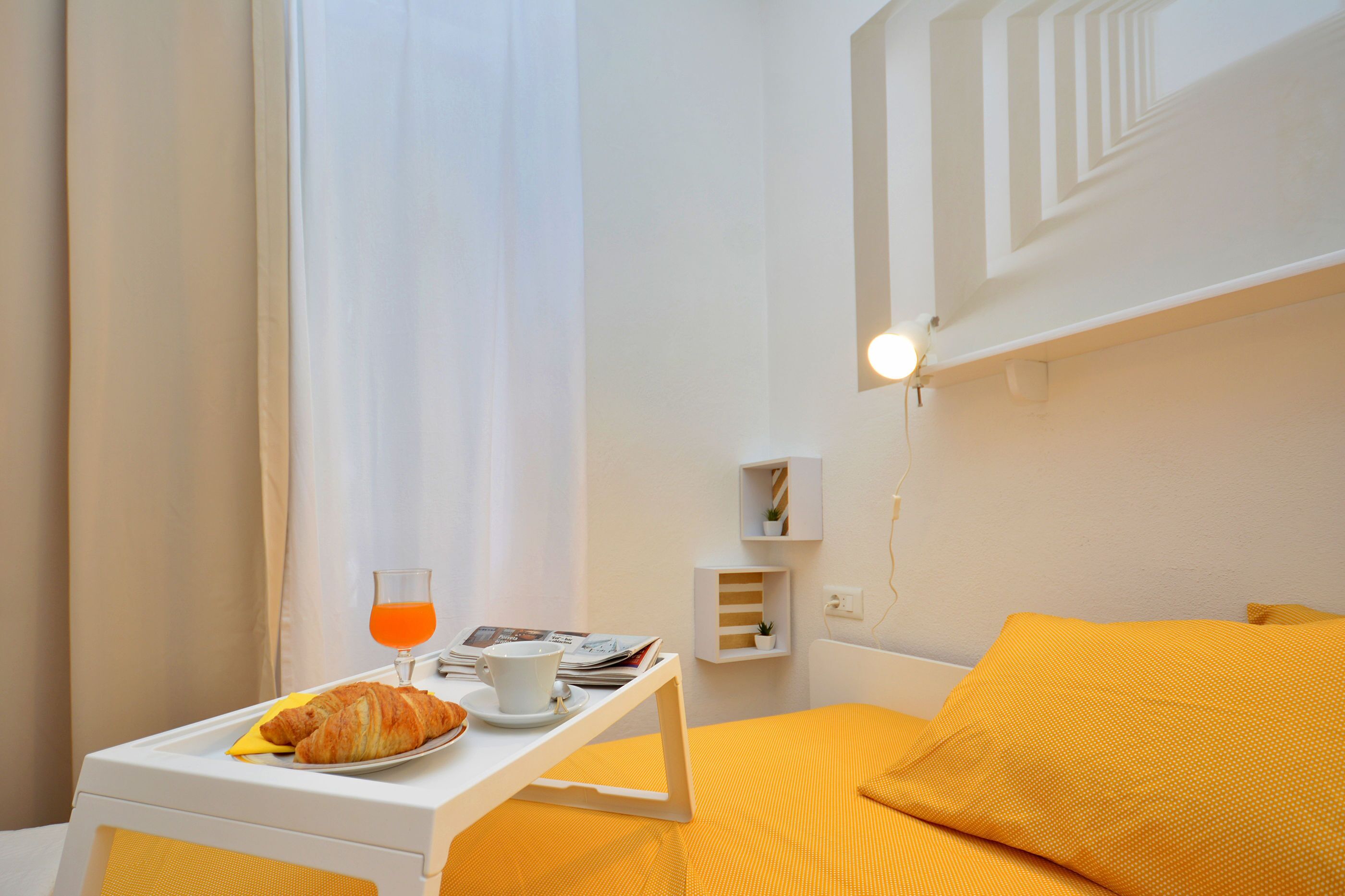 A hotel room with a bed, a bedside table with a croissant, a cup of coffee, and a glass of orange juice.