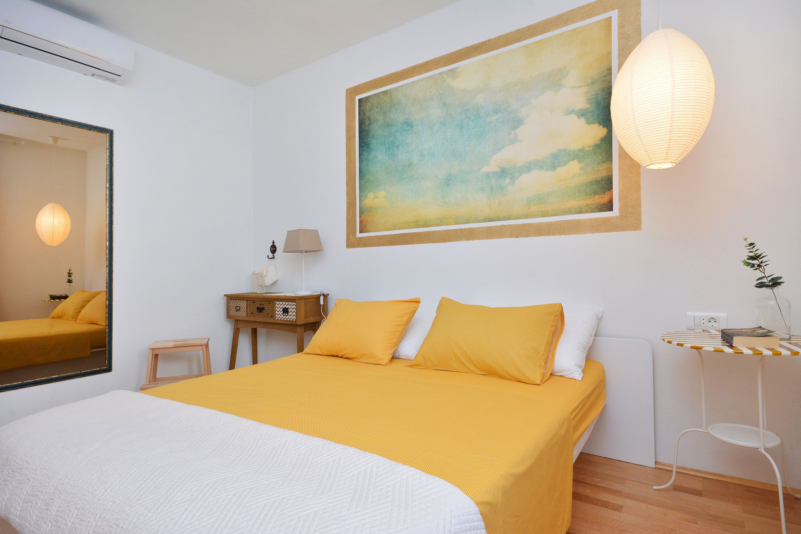 A hotel room with a bed, a bedside table, a lamp, a mirror, and a painting on the wall.
