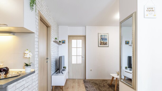 Una cocina moderna con salpicadero de ladrillo, puerta de madera y una pequeña zona de estar con televisor y objetos decorativos.