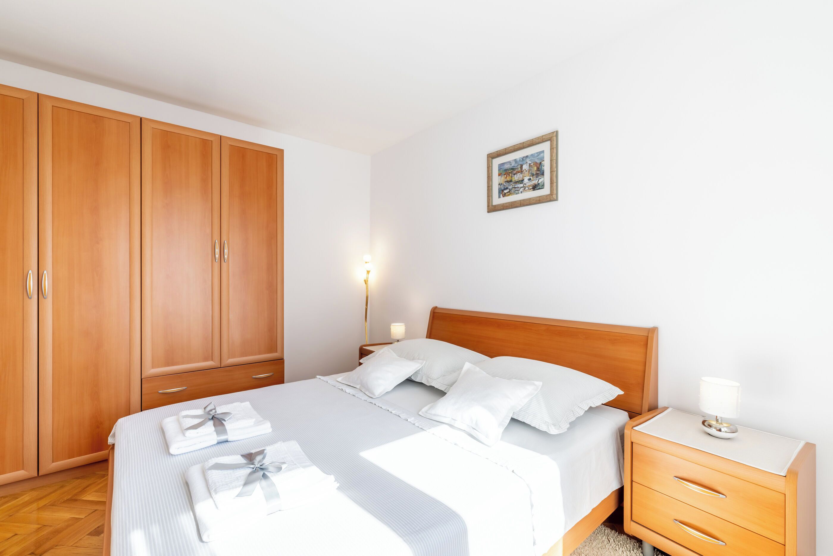 Een keurig opgemaakt bed met witte lakens en kussens, een houten kledingkast, een nachtkastje met een lamp en een lijst met een afbeelding aan de muur.